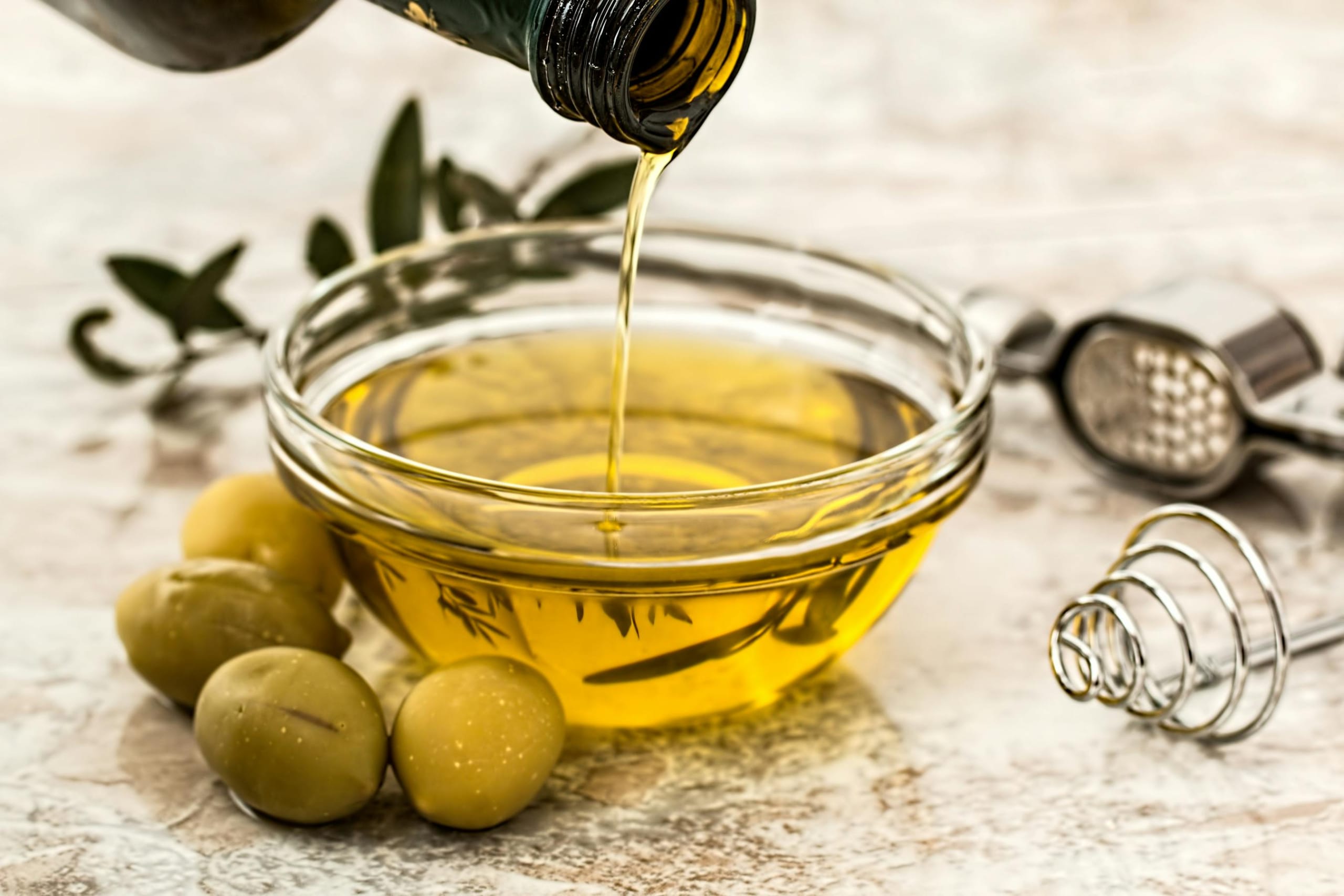 Extra virgin olive oil being poured into a glass bowl surrounded by fresh olives